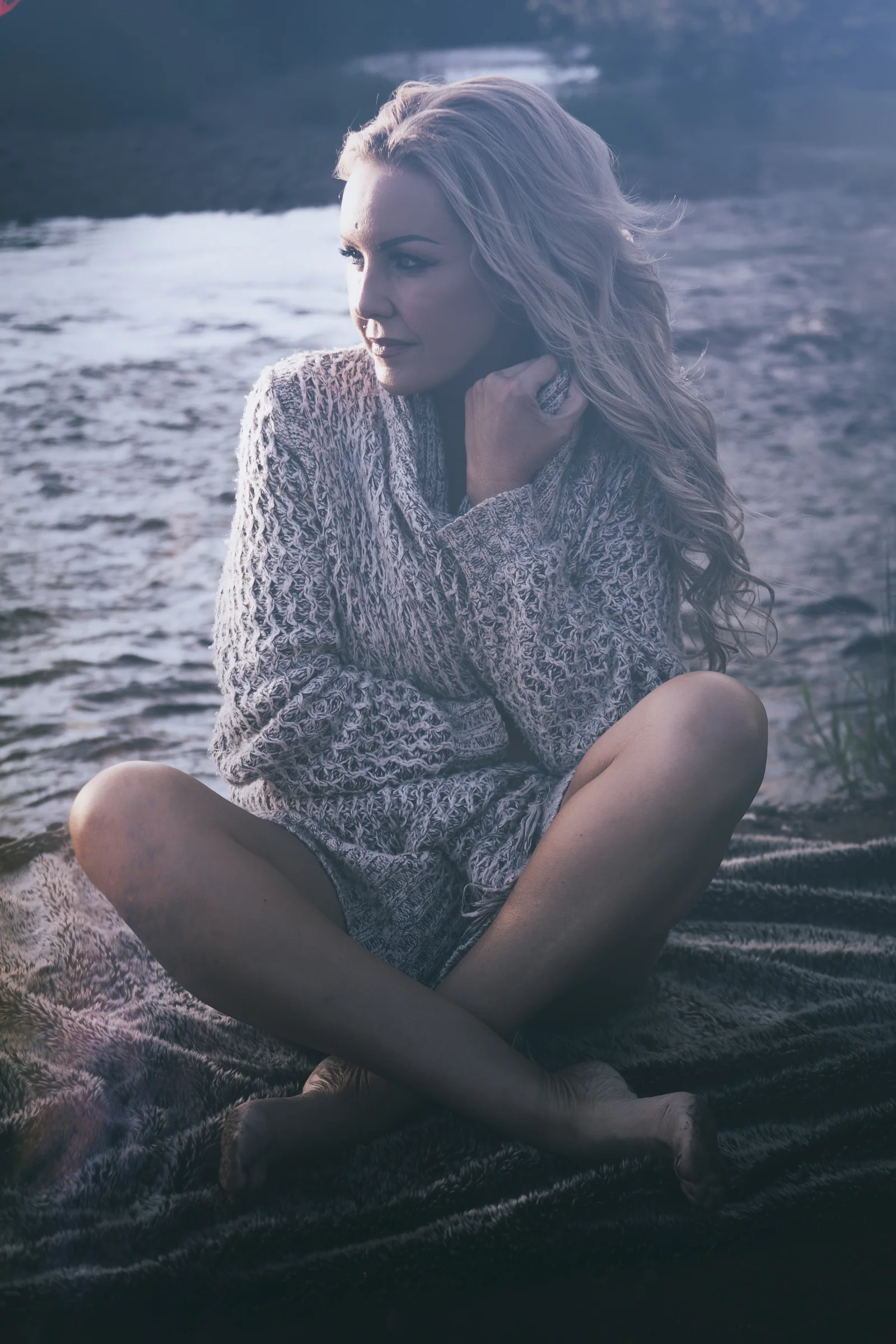A woman sits alone outdoors beside water under a muted sky.
