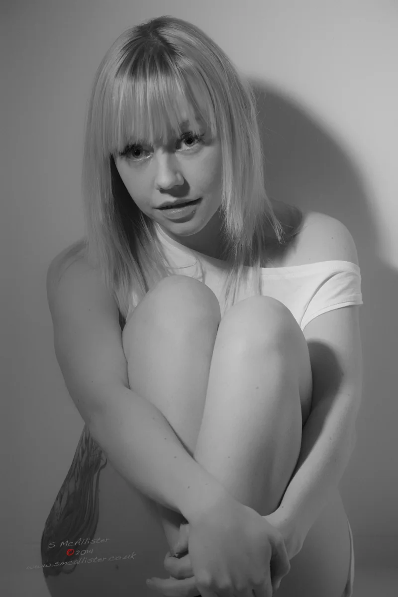 A seated woman holds her knees close in a pale studio portrait.